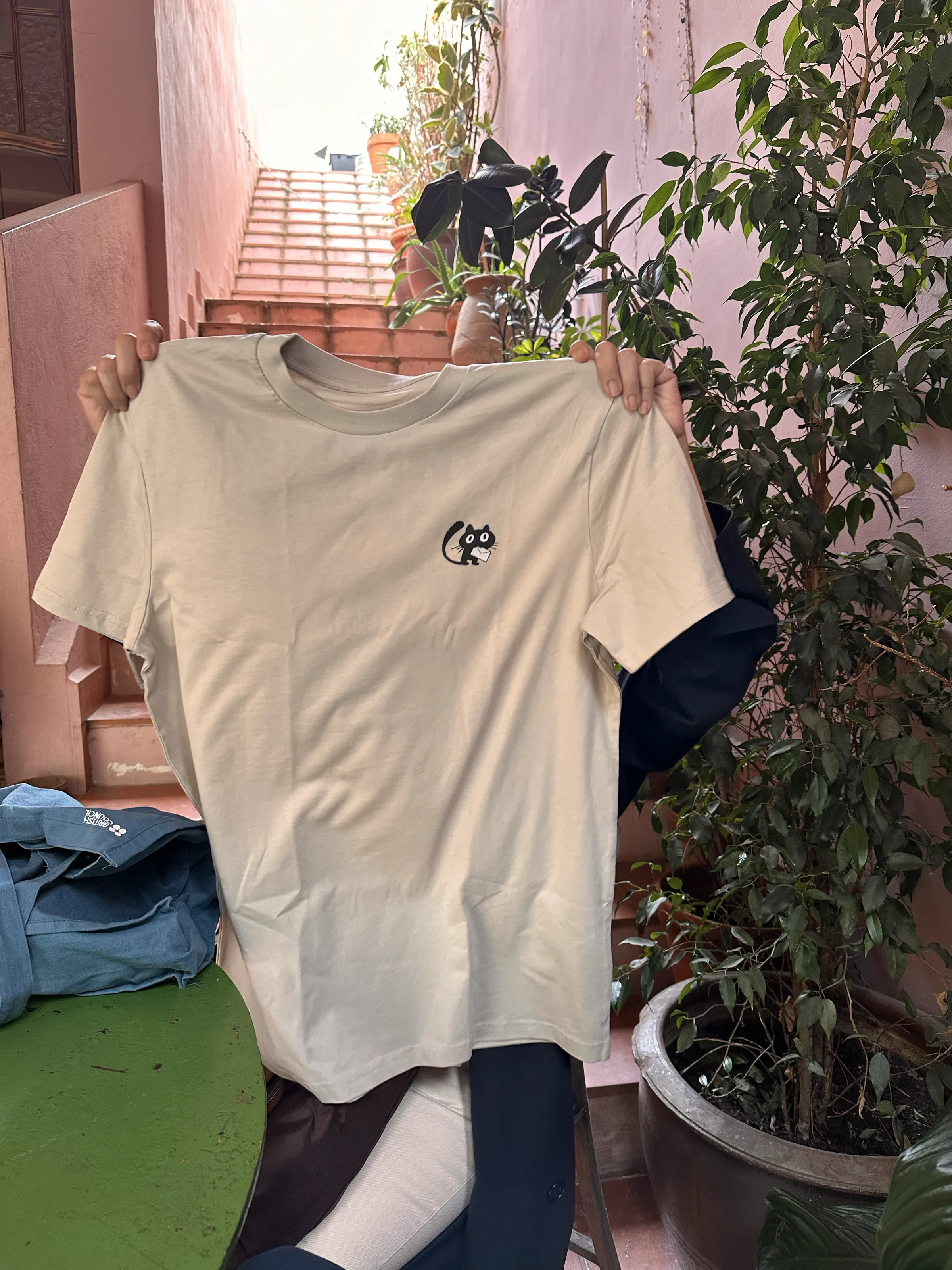 Rumicat — sand-coloured tee held up in a Barcelona stairwell, small cat embroidered on the chest